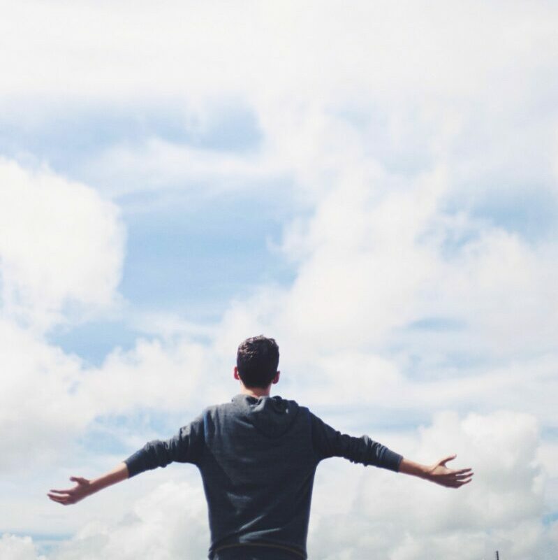From below back view of anonymous male traveler standing with reached arms enjoying cloudy sky during vacation