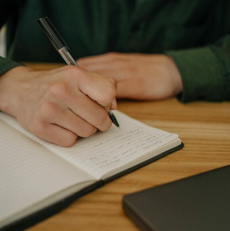 Close-up of a hand writing notes in a notebook on a wooden desk indoors.