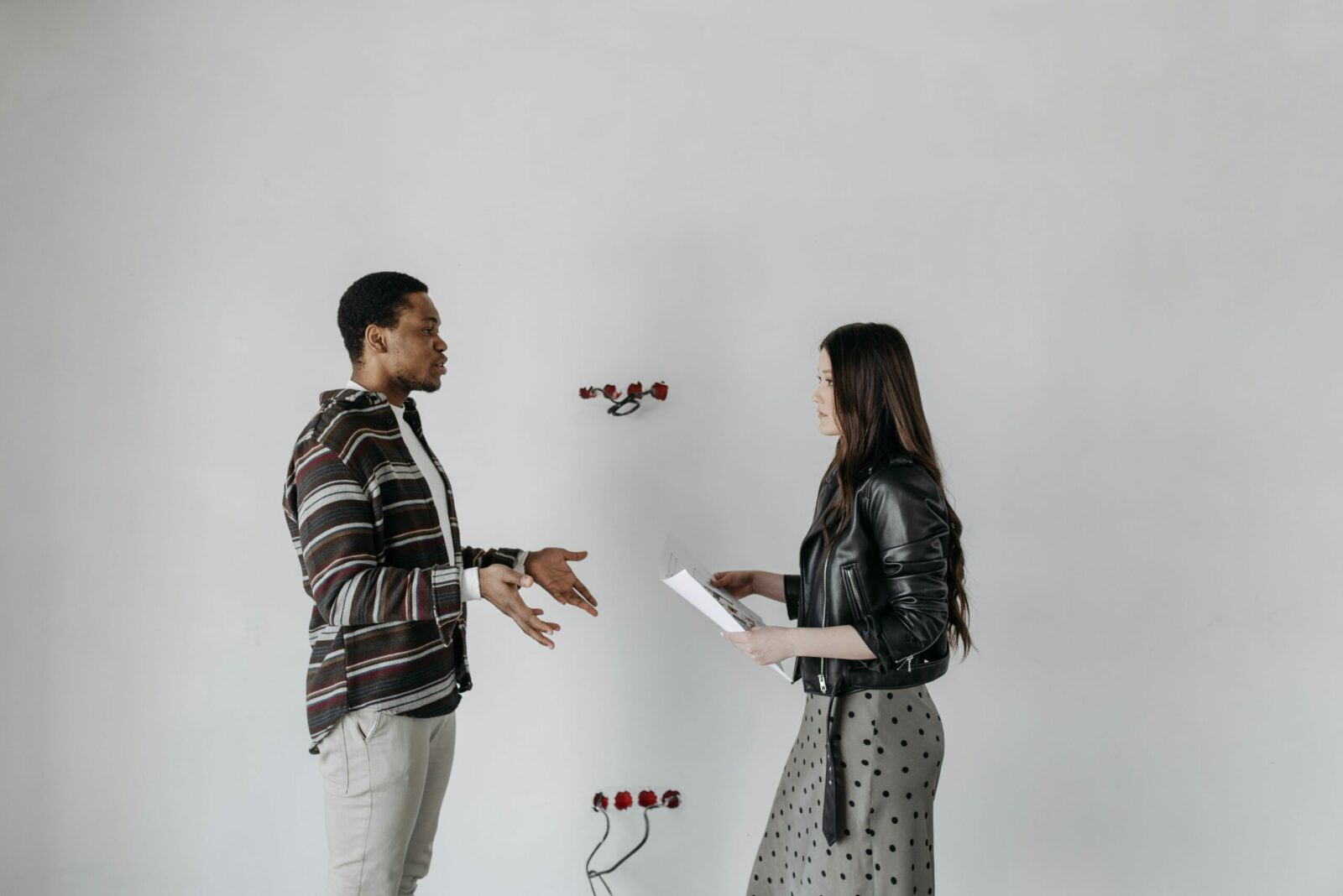 A man and woman in stylish outfits having a discussion in an interior setting with a white wall.